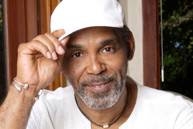 A handsome Black man with graying beard in a white hat and t-shirt smiles gently into the camera 