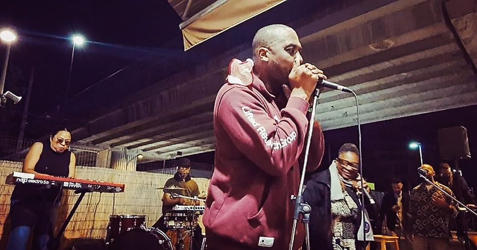 A black man with a shaved head and maroon sweatshirt performs in front of a microphone with a backing band
