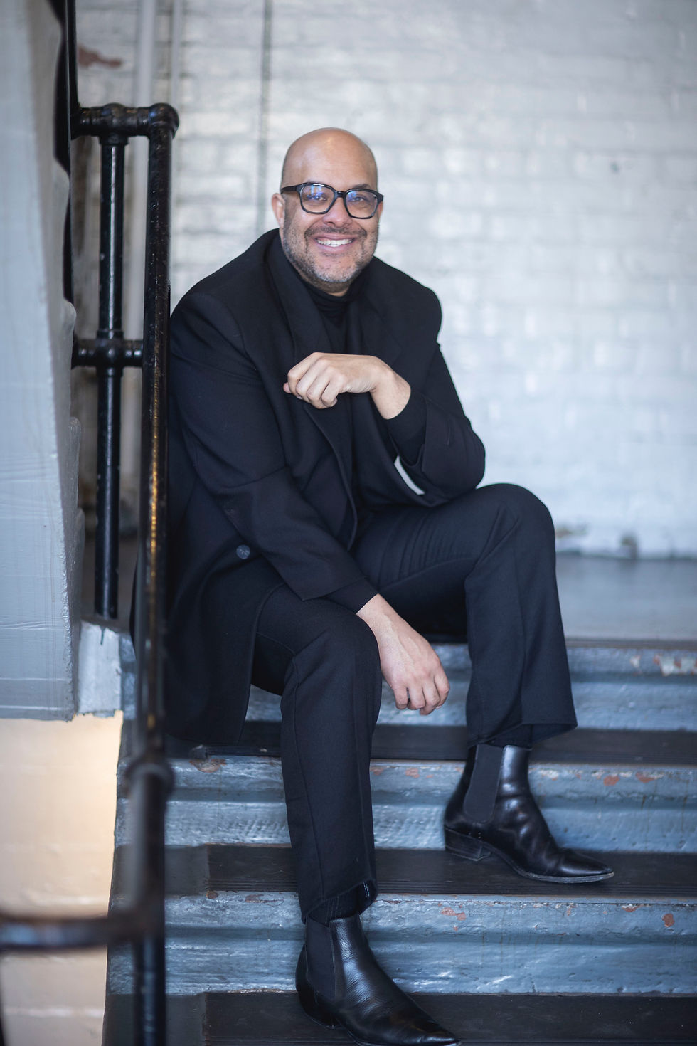 A light-skinned Black man in a tailored suit and black boots and glasses sits on a staircase and smiles into the camera.