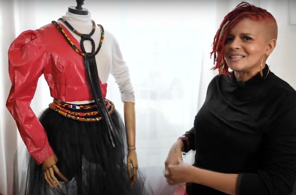 A brown skinned woman with pink dreadlocks stands next to a mannequin with colorfully designed outfit