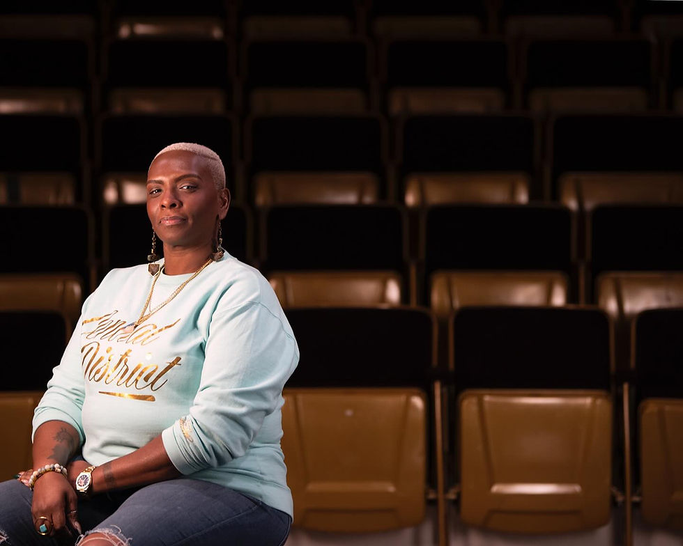 A Black woman with cropped blonde hair poses in a theatre auditorium, wearing a pale blue sweatshirt and gold jewelry