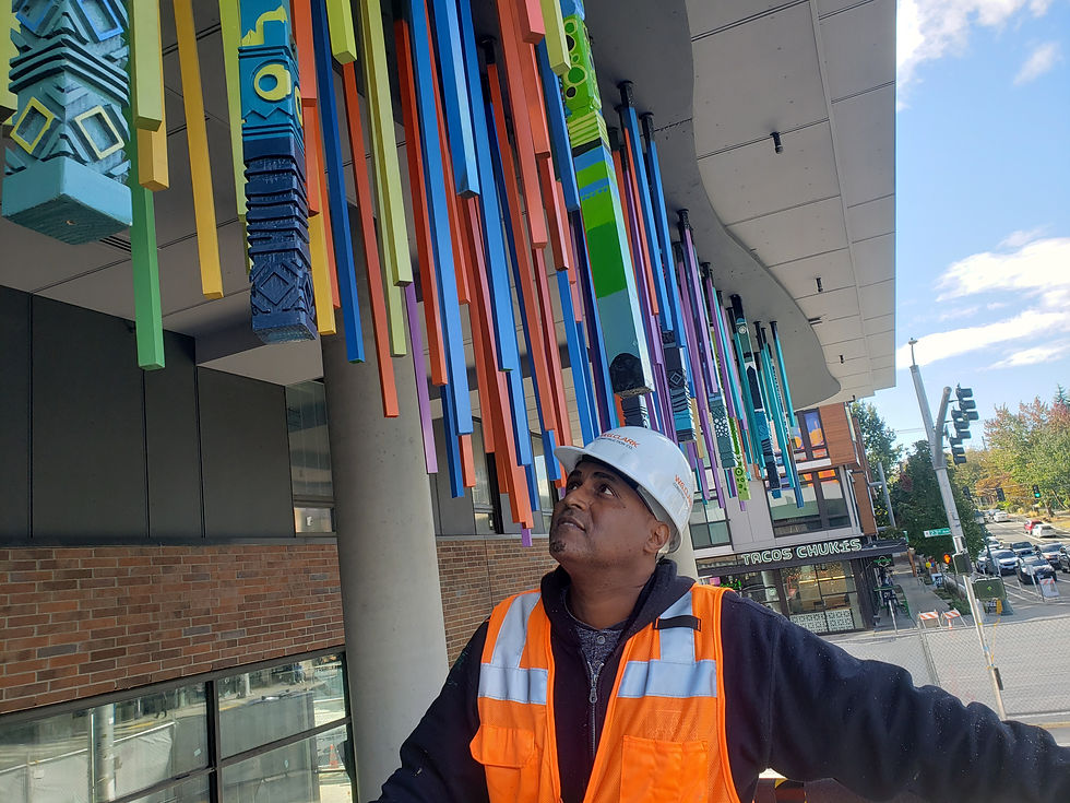 Black man in hardhat and orange reflective vest looks up at his colorful sculpture of Eritrean symbols etched on wood blocks