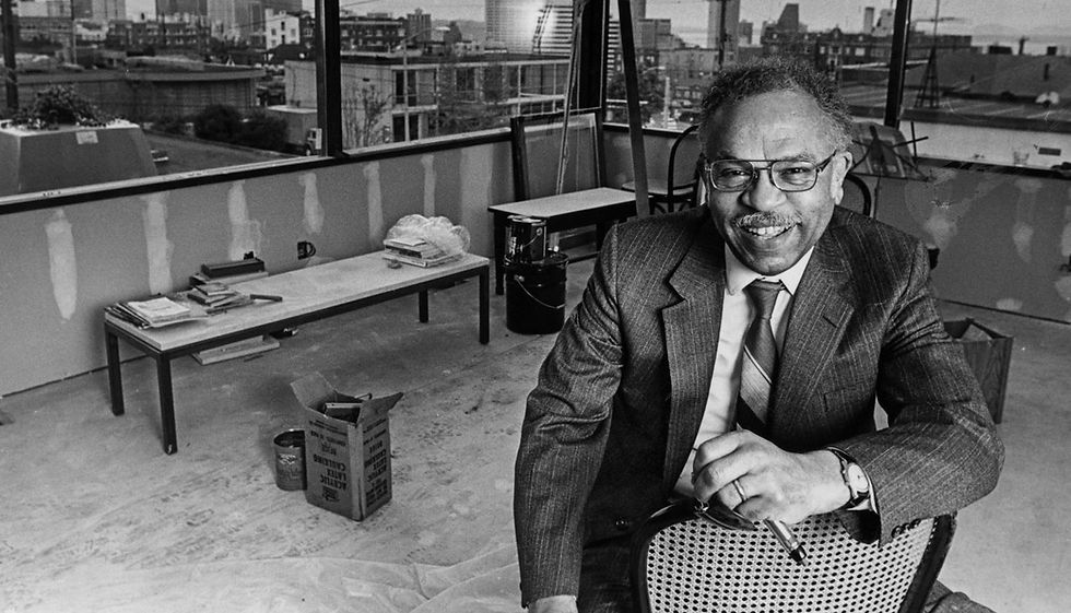 Black and white image of middle aged Black man in glasses and a suit in his architecture office