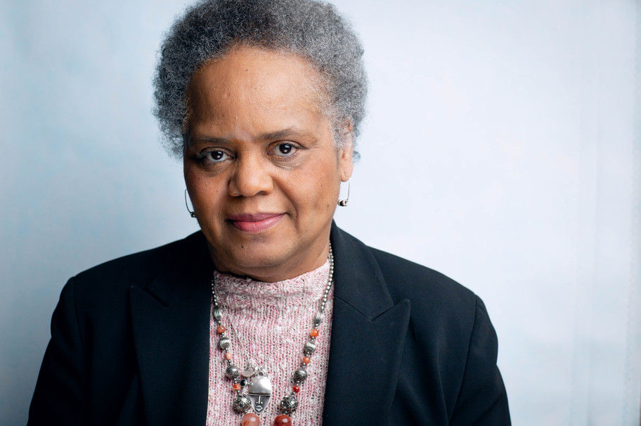 A senior Black woman with short graying hair, stylish jewelry and a light purple sweater under a black jack smiles slightly into the camera