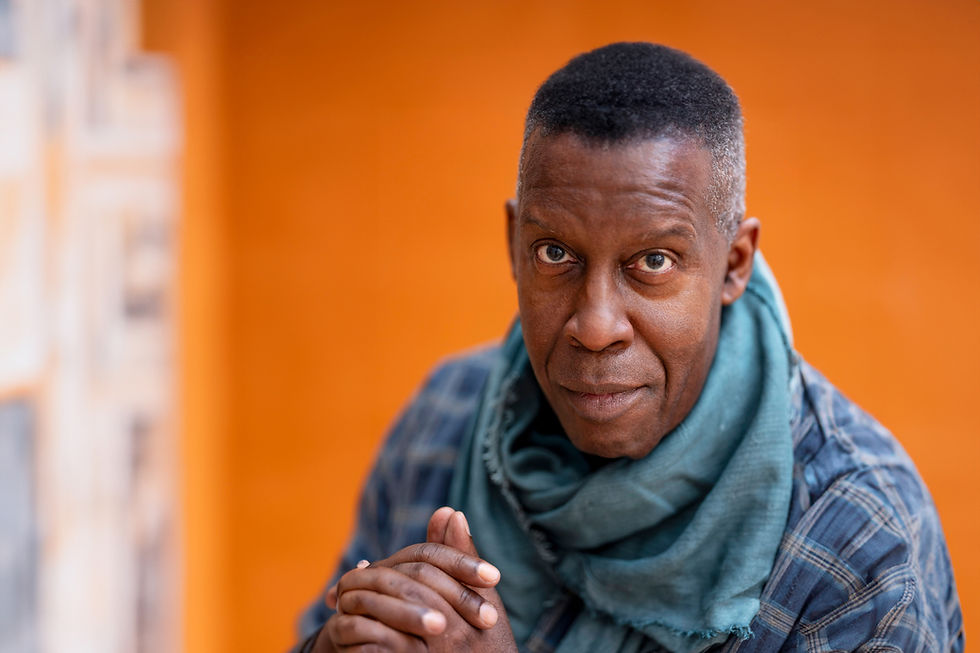 Middle-aged Black man with graying hair at temples in a turquoise scarf and flannel shirt poses in front of a vibrant orange wall