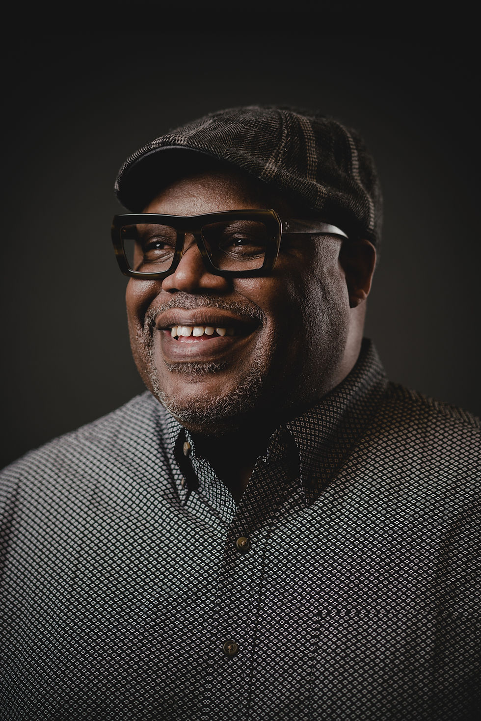Middle aged Black Man in a tweed cap, stylish black glasses and a button up shirt smiles to the left of the camera