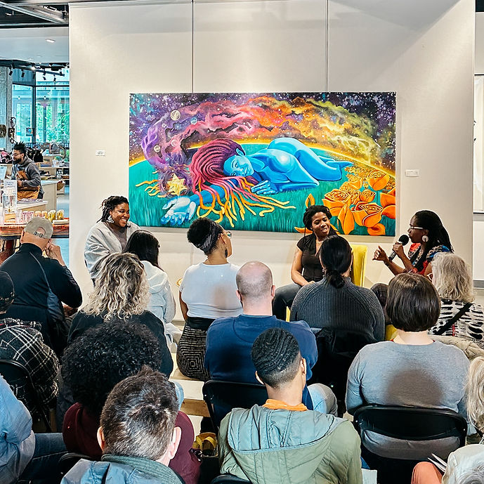 An audience sits in front of a three Black women artists and an Afro-futuristic painting in a gallery space