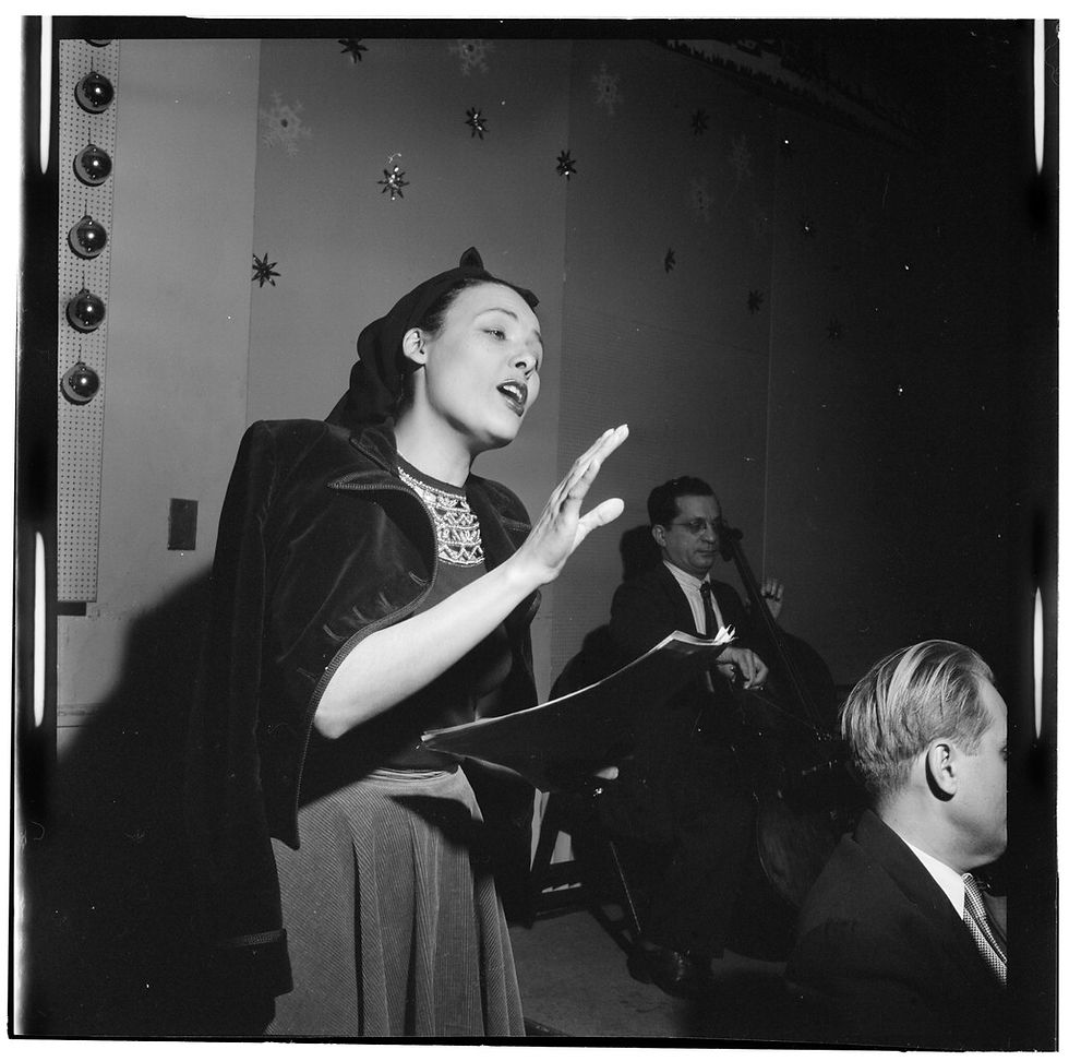 Black and White photogragph of singer and actress Lena Horne, a light-skinned black woman, holding a folder and singing with her hand raised