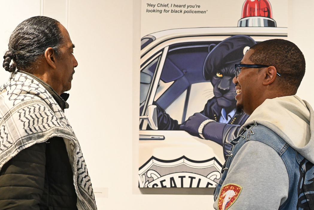 An older Black gentlemen with a scarf talks to a younger Black gentlemen in glasses and a jean jacket in front of a piece of art depicting a Black Panther driving a police vehicle. Black Panthers Co-Founder Aaron Dixon talks with artist Charles Conner about his art work
