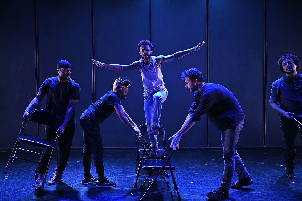 Performers under blueish light on stage dance atop and around chairs.