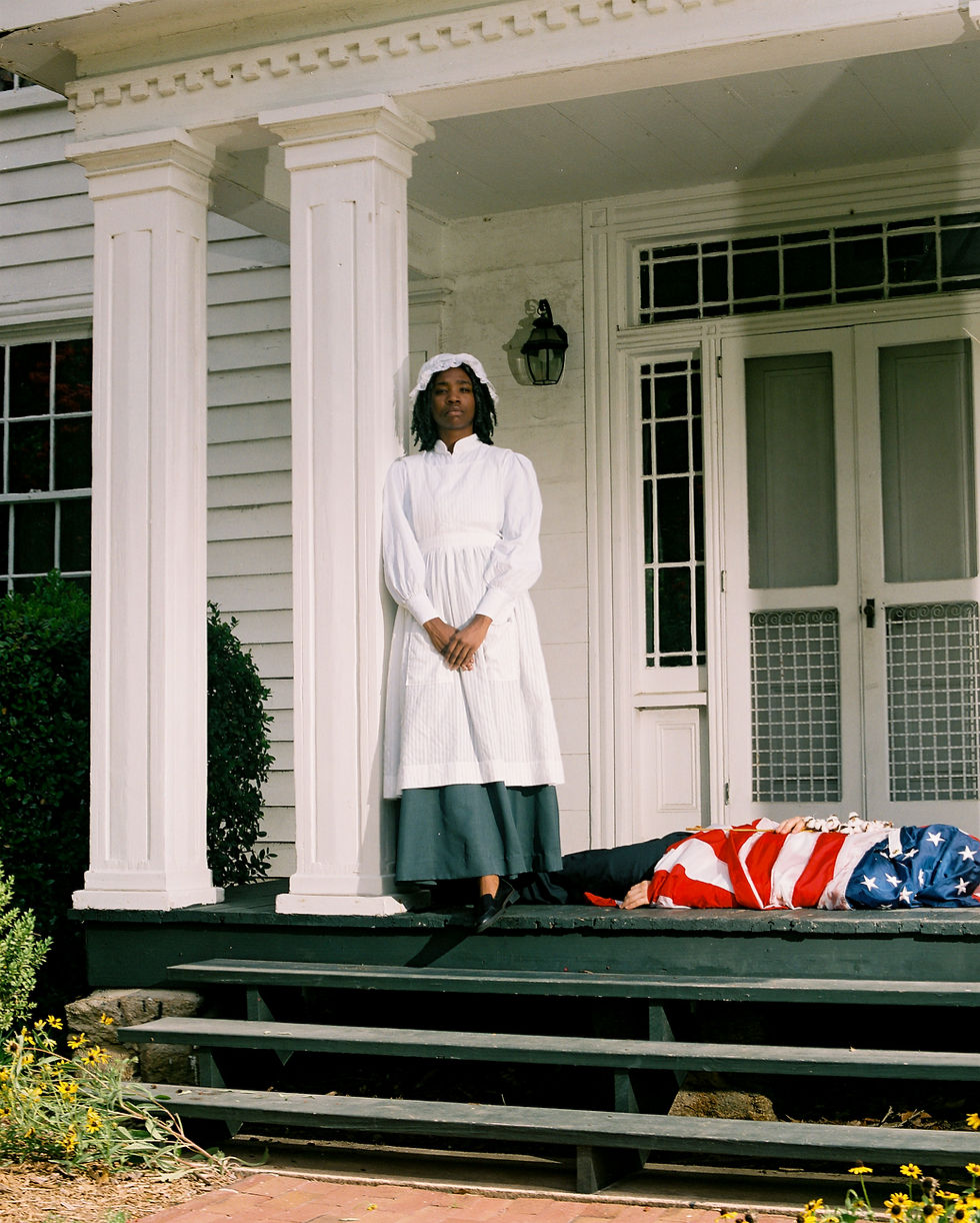 A Black woman in period clothing stand in front of a plantation style home with a person wrapped in an American flag laying at her feet.